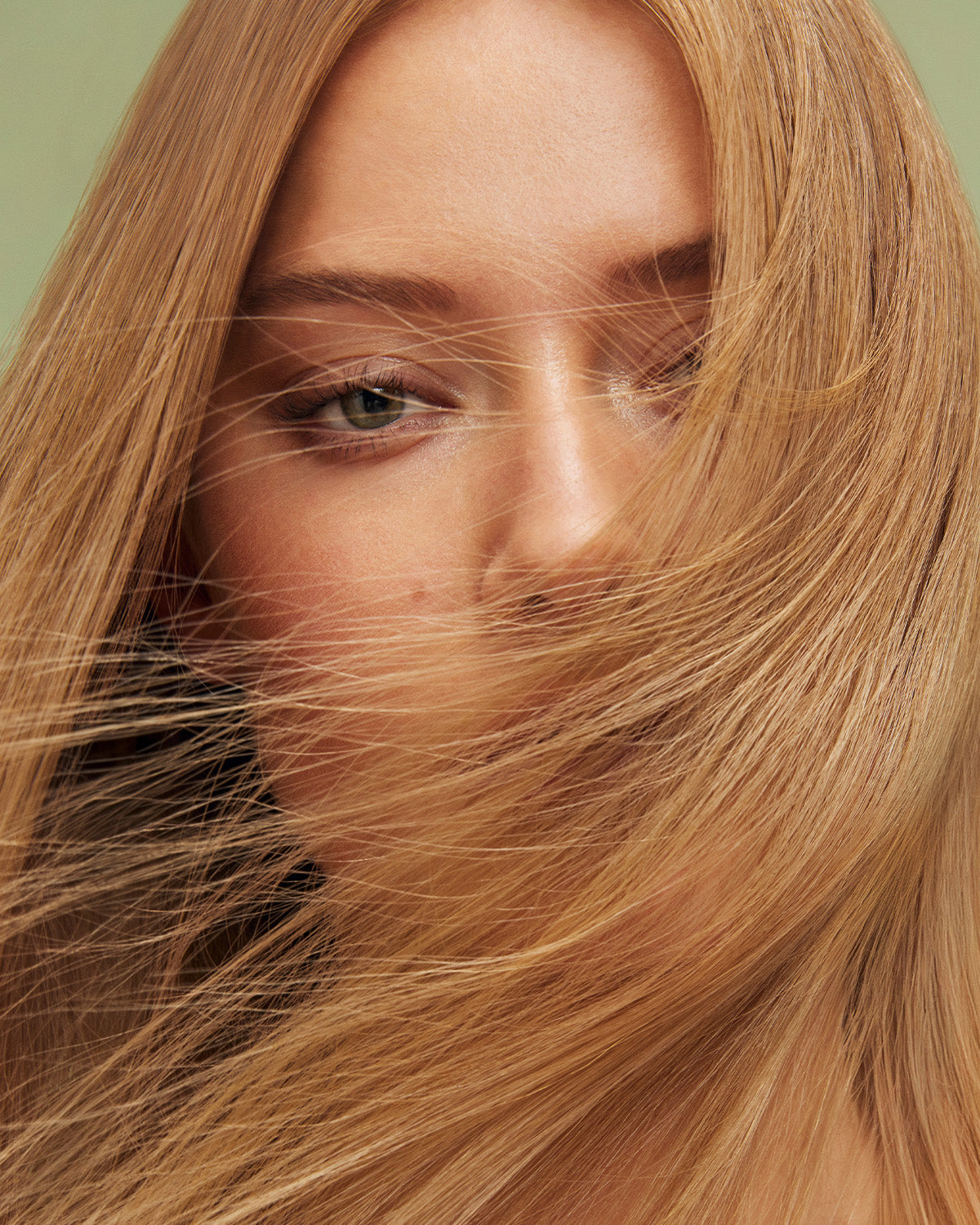 Närbild på person med långt blont hår i nyansen Beige Blonde, delvis täckt ansikte mot ljusgrön bakgrund.